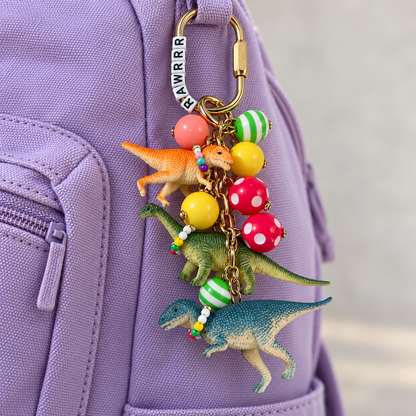 Dinosaur backpack charm with three colorful dinosaurs wearing tiny beaded necklaces, patterned resin beads, and a gold-tone clip attached to a lavender backpack.

