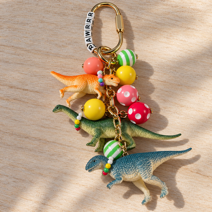 Dinosaur purse and backpack charm with three colorful dinosaurs wearing tiny necklaces, patterned resin beads, RRAWRRR letter beads, and a gold-tone clip.

