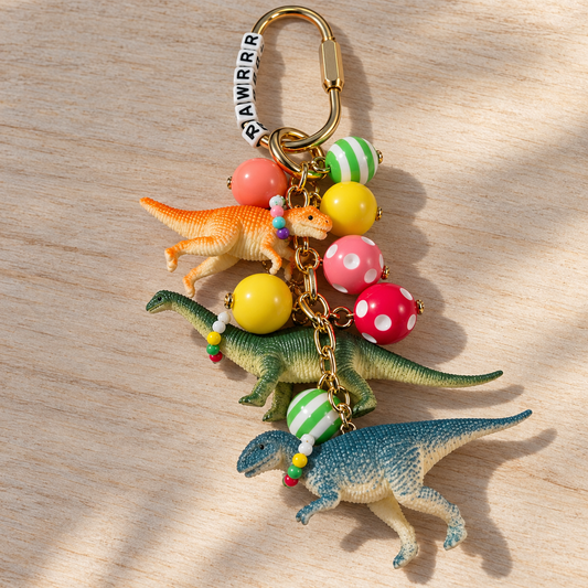Dinosaur purse and backpack charm with three colorful dinosaurs wearing tiny necklaces, patterned resin beads, RRAWRRR letter beads, and a gold-tone clip.

